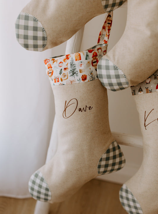 Handmade personalised Christmas stocking in natural linen with the name “Dave” in brown script font, featuring a festive Santa Claus print cuff with reindeer, Christmas trees and gifts, and finished with sage green gingham heel and toe patches. Rustic farmhouse style holiday stocking, custom made in Australia, ideal for family or children’s Christmas décor.