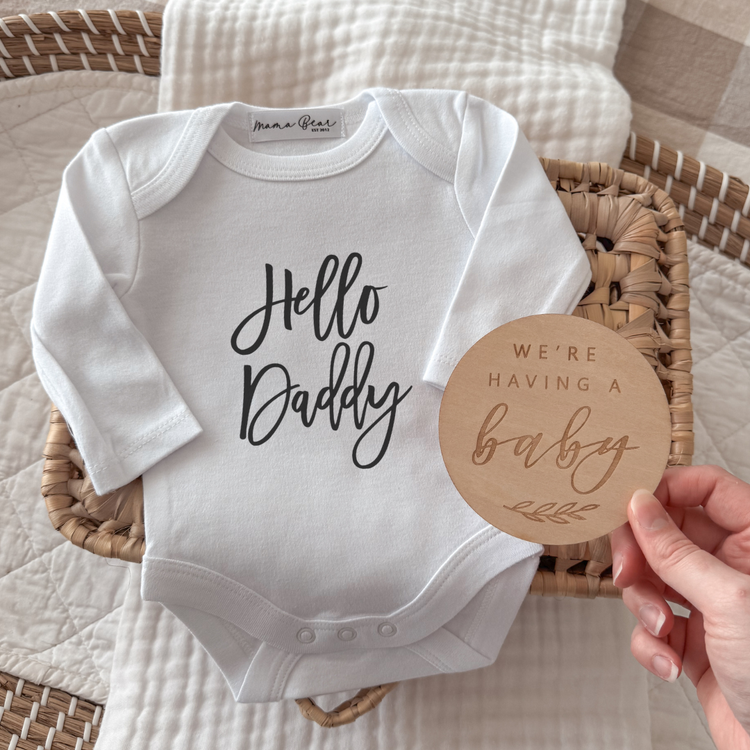 White baby onesie with 'Hello Daddy' text and a 'We're Having a Baby' coaster on a textured surface.