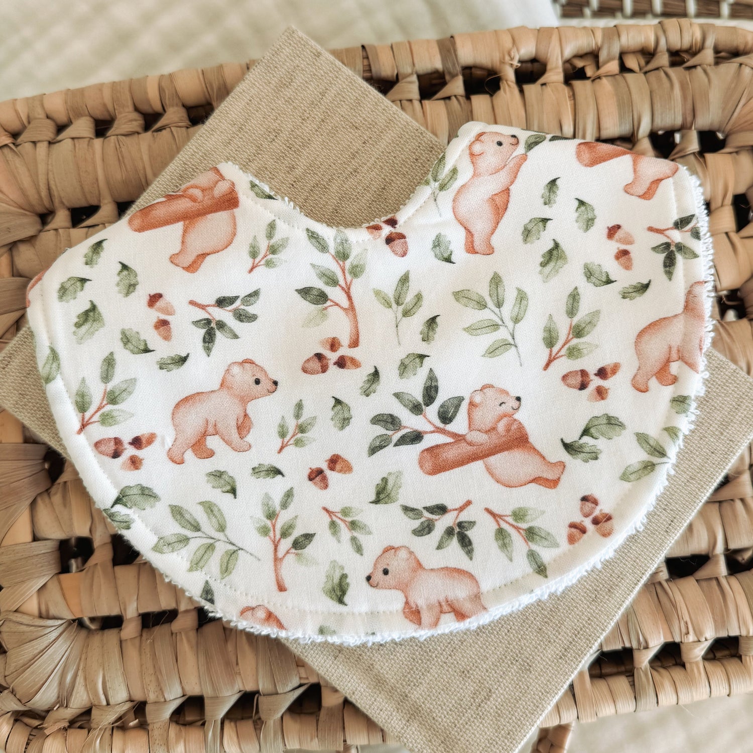 Handmade baby bib with a woodland bear print on a soft cream background. The fabric shows small cubs, leafy branches, and acorns in earthy tones. Bib is curved in shape and backed with terry towelling, laid on a neutral cloth and woven basket.
