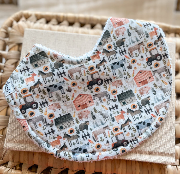 Handmade baby bib with a farmyard pattern including animals, tractors, barns, and sunflowers in muted tones; backed with absorbent terry towelling and suitable for newborns and teething babies.