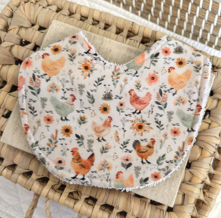 Baby bib: Background beige, illustrations of hens and wildflowers scattered. Main colours are yellows and sage green.