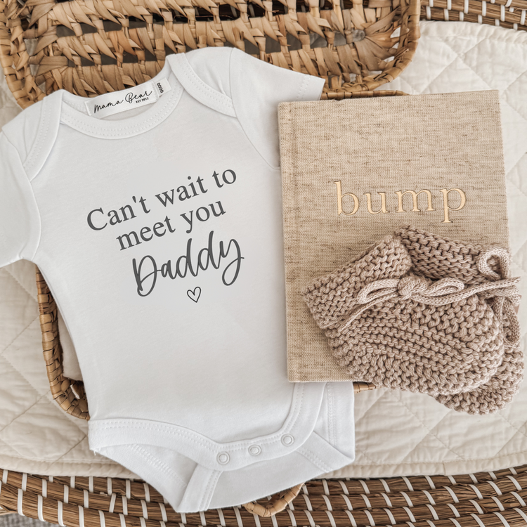 Pregnancy announcement , baby reveal to partner. White baby onesie that says in black text 'can't wait to meet you' (in times roman font), 'Daddy' (in script font) with a small dainty heart underneath. Photo props in flat lay image also include an oatmeal linen 'bump' pregnancy journal with a pair of beige booties.