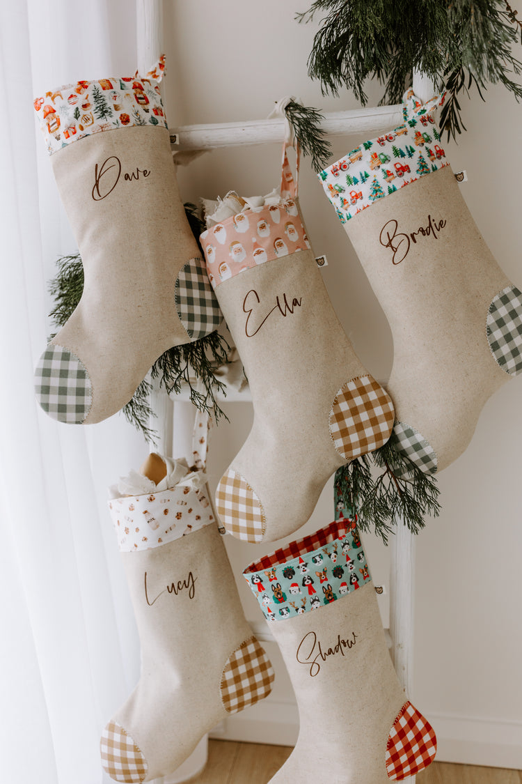 Set of five personalised Christmas stockings hanging on a rustic white ladder. Each handmade stocking is sewn from natural linen with contrasting gingham heel and toe patches, and features unique festive cuff prints including Santa Claus faces, farm tractor scenes, milk and cookies, and Christmas dogs. Stockings are customised with names in elegant script font. Perfect personalised family Christmas stockings, handmade in Australia, festive décor for kids, pets, and adult