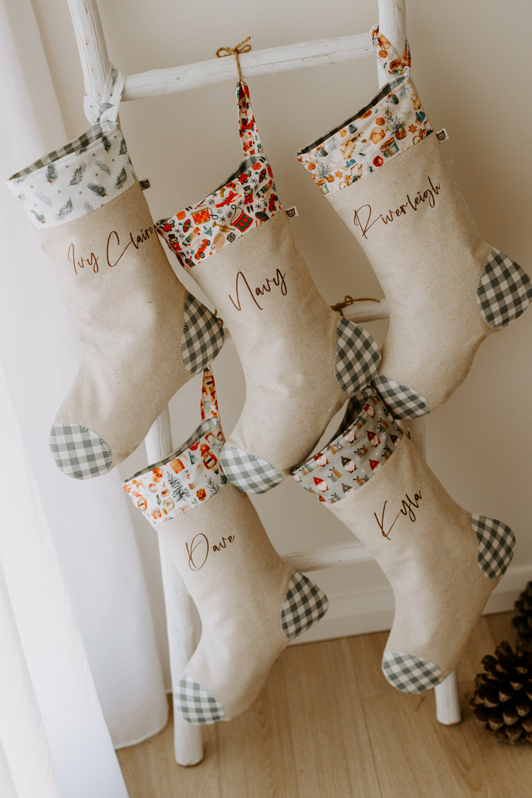 Set of family Christmas stockings coordinating fabrics, custom names Australia. Collection of five personalised Christmas stockings in natural linen with green gingham heels and toes, featuring assorted festive cuffs including pine branches, nutcrackers, reindeer and Christmas trees.