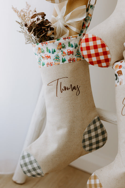 Farm-inspired personalised Christmas stocking with the name “Thomas” in script font, crafted from natural linen fabric. The stocking has a festive cuff printed with red tractors, green pine trees, and tiny holiday details, paired with a red gingham toe and muted green gingham heel patch. Styled on a rustic white ladder with wrapped gifts and native dried foliage, the scene captures a warm, country farmhouse Christmas aesthetic.