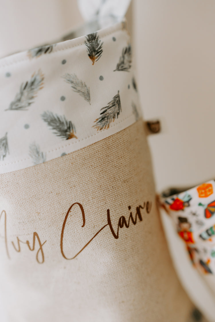 Close up of Christmas stocking, beige stocking with name Ivy Claire in brown script don’t. Trim of stocking is white with scattered pine leaf branches, making for a gender neutral stocking.