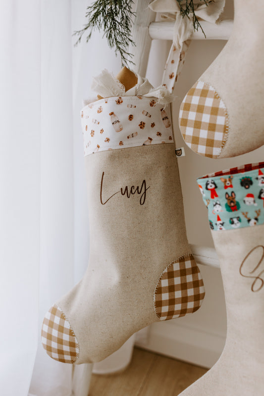 Personalised Christmas stocking with the name “Lucy” in script font, handmade from natural linen and finished with caramel gingham patchwork heel and toe. The cuff features a sweet white fabric decorated with festive gingerbread men and candy canes, creating a warm, nostalgic holiday look. Styled hanging on a white ladder with sprigs of greenery for a cosy farmhouse-inspired Christmas display.