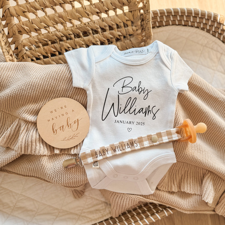 White baby bodysuit featuring personalised 'Baby (Surname) Williams' (Script font) JANUARY 2025 (due Date in times roman font) for a surprise baby announcement. A wooden disc saying 'we're having a baby' and a personalised caramel gingham dummy clip saying 'BABY WILLIAMS' on it.