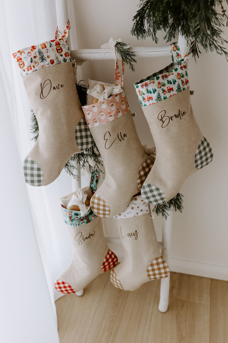 Set of six personalised Christmas stockings hanging from a rustic white wooden ladder, each made from natural linen with gingham heels and toes. The stockings feature a mix of festive cuff fabrics: red tractor farm print, peach Santa faces, puppy dogs in Christmas accessories, woodland animals, and vintage-inspired toy designs. Names including Dave, Ella, Brodie, Shadow, and Lucy are printed in brown script font across the fronts. Styled with sprigs of greenery and wrapped gifts, the scene captures a warm,