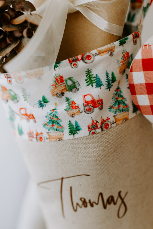 Close-up of a personalised Christmas stocking made from natural linen, featuring the name “Thomas” in brown script font. The cuff is decorated with a farm-themed festive print of bright red tractors, green pine trees, and little Christmas details like presents and decorated trees. The stocking is styled with rustic touches, including wrapped gifts tied with sheer white ribbon and dried native foliage, creating a warm farmhouse Christmas vibe.