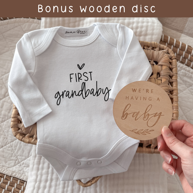 White baby onesie with 'first grandbaby' text and a wooden disc reading 'We're having a baby' on a textured surface.