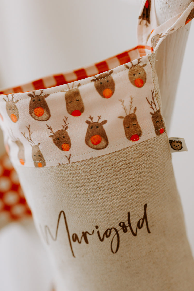 Close-up of handmade personalised Christmas stocking in beige fabric with red gingham trim and reindeer print cuff, customised with name Marigold.