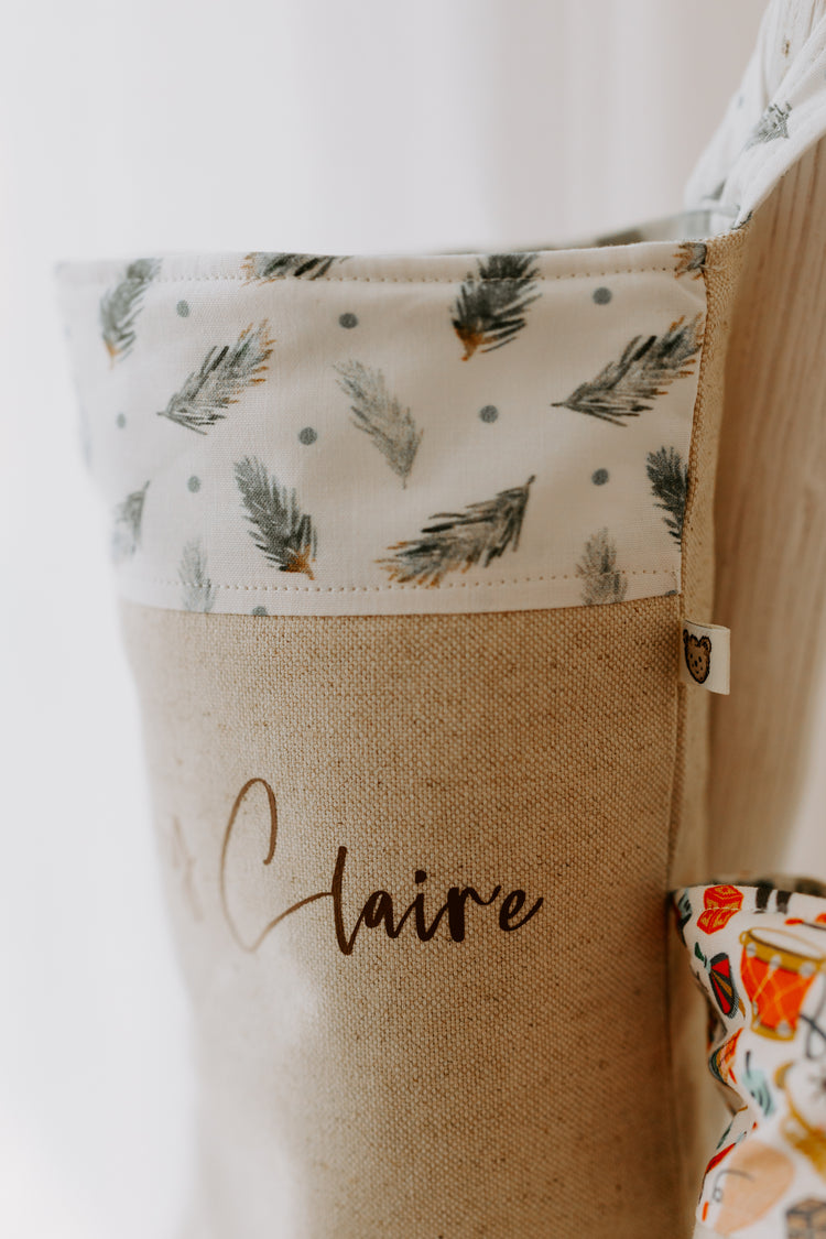 Baby first Christmas stocking with soft green leaves, personalised with name. The stocking is made of a beige linen type fabric, with the Ivy Claire written in brown script font the trim of the stocking is white with illustrations of green pine leaves.