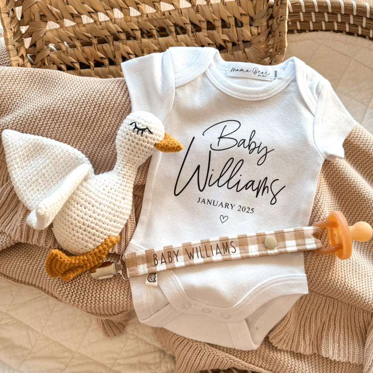 Social media flat lay of a personalised white baby onesie with custom surname and due date. Perfect for a pregnancy reveal to family and friends. Onesie is laying in a wicker basket with a beige bay blanket. A short sleeve onesie that says 'Baby Williams' in Script font followed in small times roman font 'JANUARY 2025'. There is also some props in the image, a white knitted baby goose and a caramel gingham check dummy clip, personalised in brown font 'BABY WILLIAMS'.
