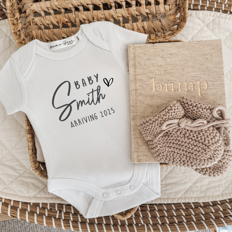 Flat lay of a white baby onesie with “Baby Smith Arriving 2025” printed in black, featuring a bold script font with a heart detail. The onesie is styled in a woven basket with a beige “bump” pregnancy journal and a pair of knitted baby booties. A modern and minimal personalised baby announcement idea, perfect for revealing your pregnancy to family or capturing milestone keepsake photos.