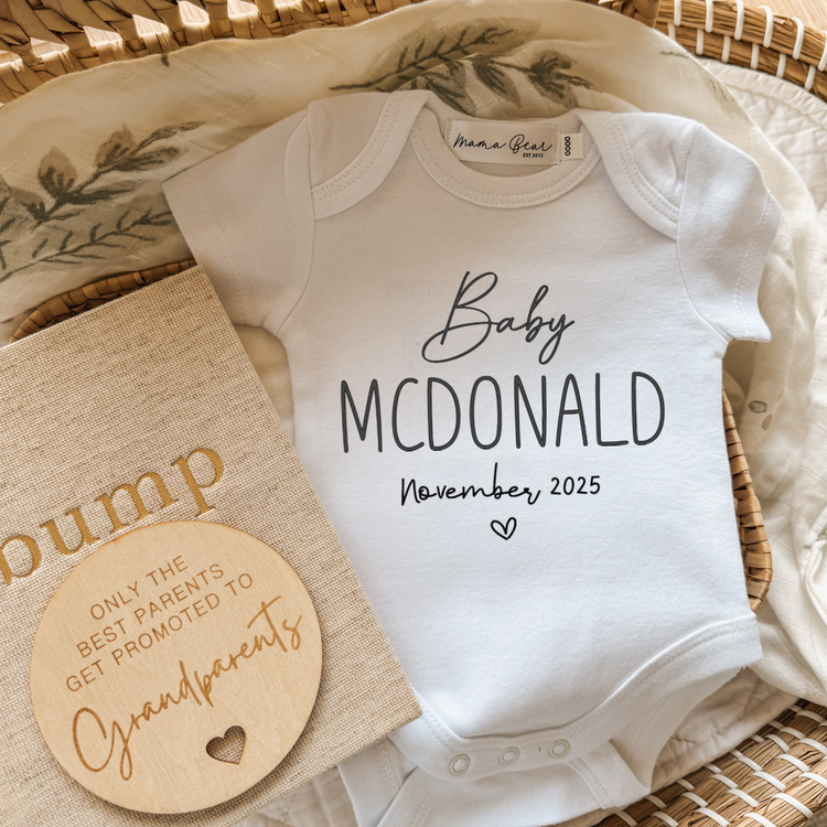 White personalised baby announcement onesie with ‘Baby JONES Coming Soon’ placed in a neutral-toned setting with a wooden disc that says ‘We’re having a baby’ and a beige linen pregnancy journal. A soft and simple pregnancy reveal outfit made in Australia.