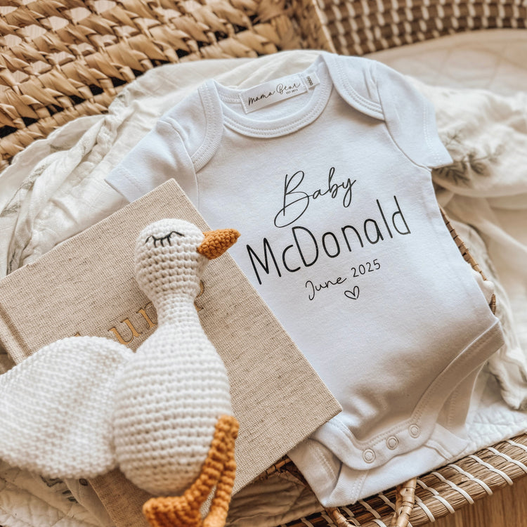 Flat lay of a personalised white baby announcement onesie with ‘Baby McDonald June 2025’ printed in a soft standard font, styled with a beige ‘bump’ journal and crochet duck toy — a sweet way to reveal a pregnancy to family or in-laws.