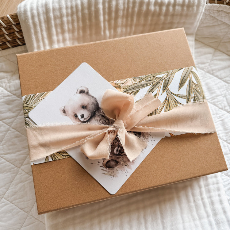 Discreet packaging for this gorgeous baby reveal surprise inside. Picture shows a square brown kraft box. With a band of greenery wrapping paper around it, tied together with a beige chiffon ribbon and tucked inside is a teddy bear postcard illustration.