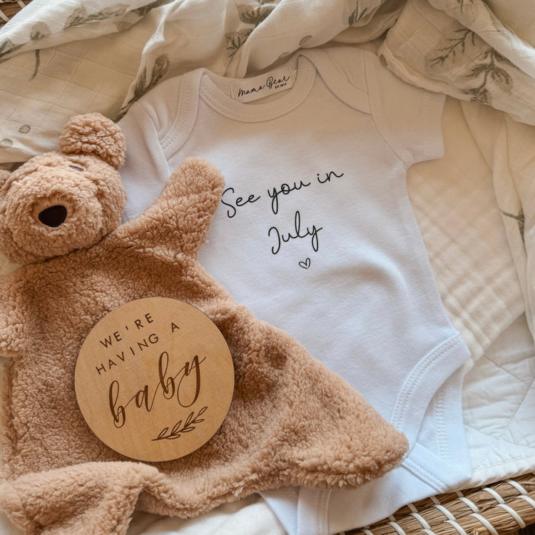 Flat lay of a white baby onesie with the text “See you in July” printed in script font with a heart underneath—perfect for a pregnancy reveal. The onesie is styled in a wicker basket on a leafy print baby wrap, paired with a soft brown teddy lovey and a wooden announcement disc that reads “We’re having a baby.” A sentimental and neutral-toned baby reveal idea for social media or gifting.