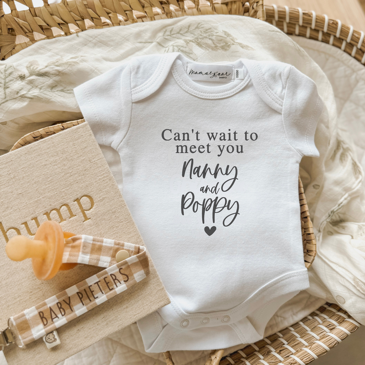 Flat lay of a white baby onesie printed with the message “Can’t wait to meet you Nanny and Poppy” in a mix of serif and script fonts. The onesie is styled inside a wicker basket on a soft botanical muslin wrap, with a beige “bump” pregnancy journal and a gingham dummy clip personalised with “BABY PIETERS.” A sentimental and customisable baby announcement idea for grandparents or in-laws.