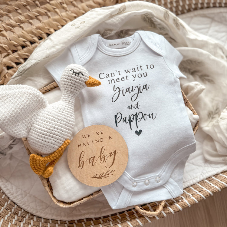 Flat lay of a personalised white baby onesie printed with “Can’t wait to meet you Yiayia and Pappou” in a mix of serif and script fonts with a small heart underneath. The onesie is styled inside a wicker basket on a leafy muslin wrap, paired with a white knitted goose toy and a round wooden plaque that reads “We’re having a baby.” A beautiful and customisable pregnancy announcement idea for announcing to grandparents, including in-laws or family members with cultural names.