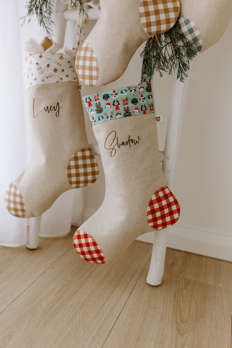 Pair of personalised Christmas stockings hanging on a white wooden ladder. One stocking reads “Lucy” with a neutral gingham trim, while the other says “Shadow” with a festive cuff featuring colourful cartoon dog faces in Santa hats, scarves, and wreaths. Both stockings are crafted from natural linen with red or tan gingham heel and toe patches. A perfect Christmas decoration for pet lovers, fur babies, and families wanting to include their dogs in holiday traditions.