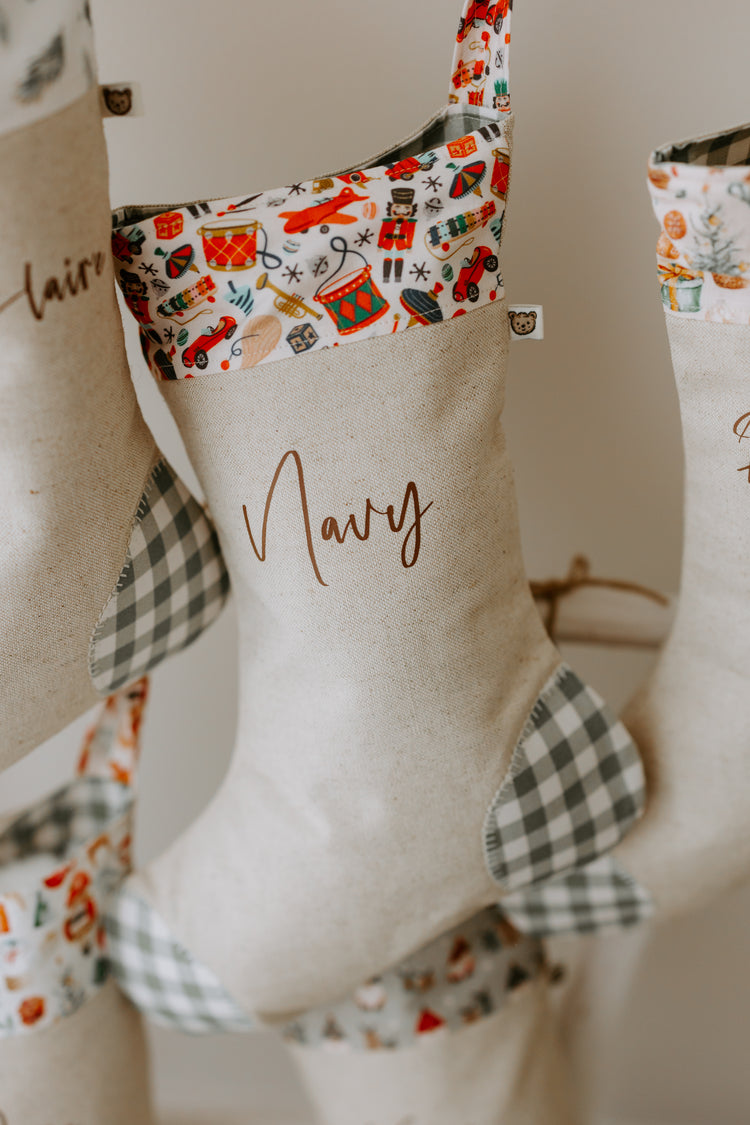 Personalised Christmas stocking in natural beige linen with the name “Navy” in elegant script. The cuff showcases a bright vintage-style Christmas toy print with nutcrackers, toy drums, rocking horses, and Santa figures in festive red and green tones. Grey gingham fabric is used on the heel and toe patches, giving the stocking a timeless handmade look that suits both kids and family holiday décor.