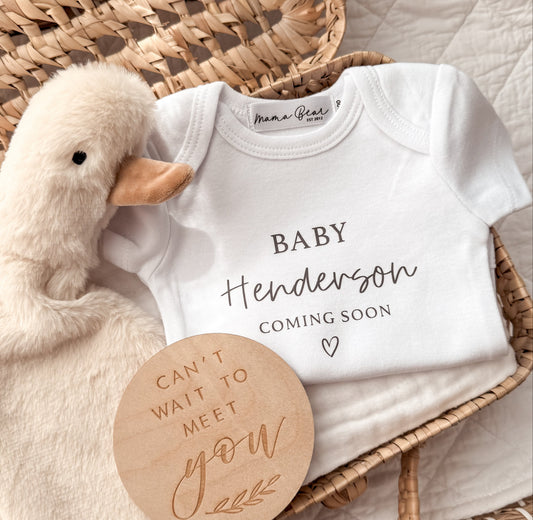 Flat lay of a white personalised baby onesie printed with the words “Baby Henderson” in script font and “COMING SOON” underneath in serif font, with a small heart icon. The onesie is styled in a wicker basket beside a fluffy white plush duck and a round wooden disc engraved with the words “Can’t wait to meet you.” A soft, neutral-toned baby announcement setup, perfect for a pregnancy reveal gift or social media post.