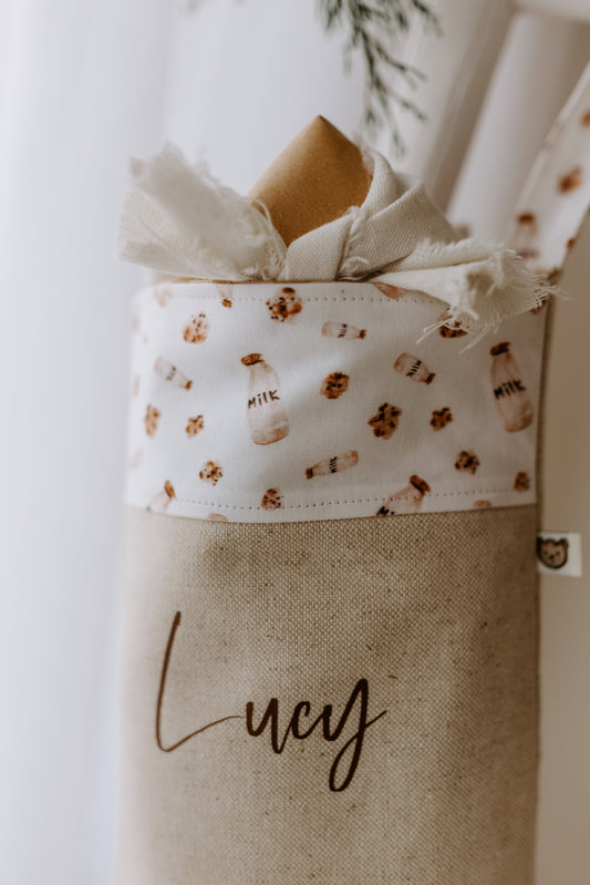 Close-up of a personalised Christmas stocking with the name “Lucy” in elegant script font on natural linen fabric. The cuff is decorated with a playful festive print featuring milk bottles and chocolate chip cookies on a white background, reminiscent of Santa’s traditional Christmas Eve treats. The stocking is styled with rustic frayed-edge fabric and greenery peeking out, adding a cosy farmhouse touch.
