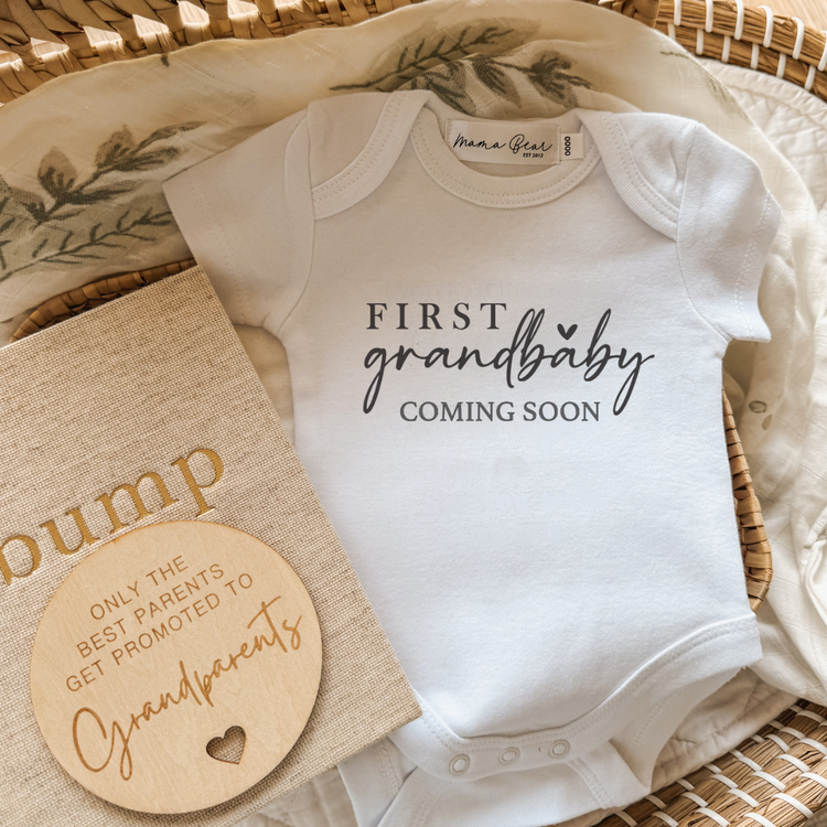 Flat lay of a white baby onesie with “First Grandbaby Coming Soon” printed in black serif and script fonts with a small heart accent. Styled in a wicker basket with a leafy muslin wrap, beige “bump” pregnancy journal, and a round wooden plaque engraved with “Only the best parents get promoted to grandparents.” A sentimental pregnancy announcement idea for first-time grandparents, perfect for gift boxes or social media reveals.