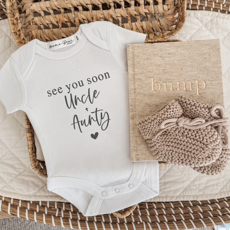 Flat lay of a white baby onesie printed with “See you soon Uncle + Aunty” in black script and serif fonts, with a small heart underneath. The onesie is styled in a wicker basket with a beige “bump” pregnancy journal and a pair of soft knitted baby booties. A customisable baby announcement onesie, perfect for sharing the news with loved ones like grandparents, aunties, uncles, or personalised names.