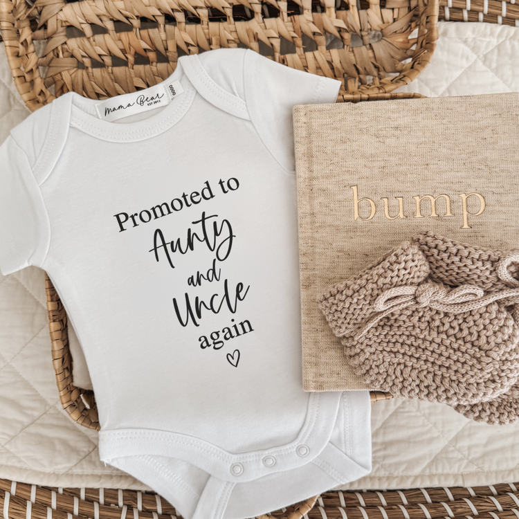 A white short-sleeve baby bodysuit with the text 'Promoted to Aunty and Uncle again' in black script, styled in a wicker basket with a beige quilt and knitted beige booties.