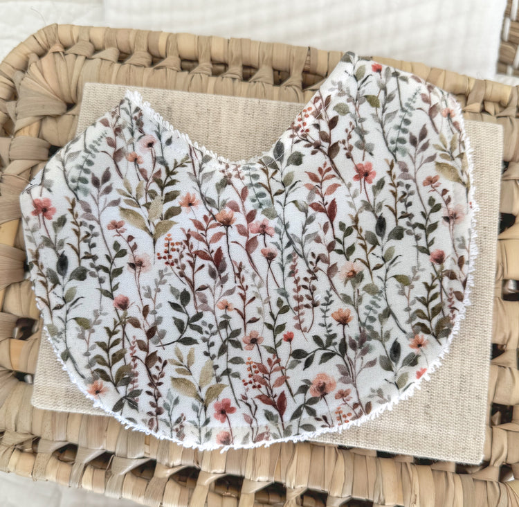 Baby bib with delicate wildflower and leaf pattern in muted blush, rust, olive, and soft brown tones on cream cotton, handmade in a contoured shape for newborn comfort.