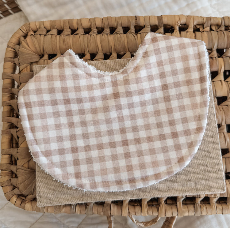 Neutral-toned handmade baby bib in a soft stone gingham print, placed on a beige linen cloth over a woven basket. The bib features curved edges and terry towelling backing, ideal for teething and dribble.