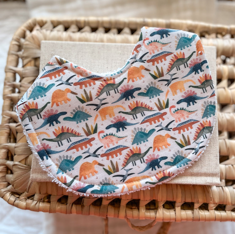 Handmade baby bib with a colourful dinosaur print in shades of teal, burnt orange, mustard, and soft grey on a cream background. Features a mix of stegosaurus and other dinosaur shapes with little leaves scattered throughout. Curved shape with terry towelling backing, photographed on a linen cloth and woven basket.