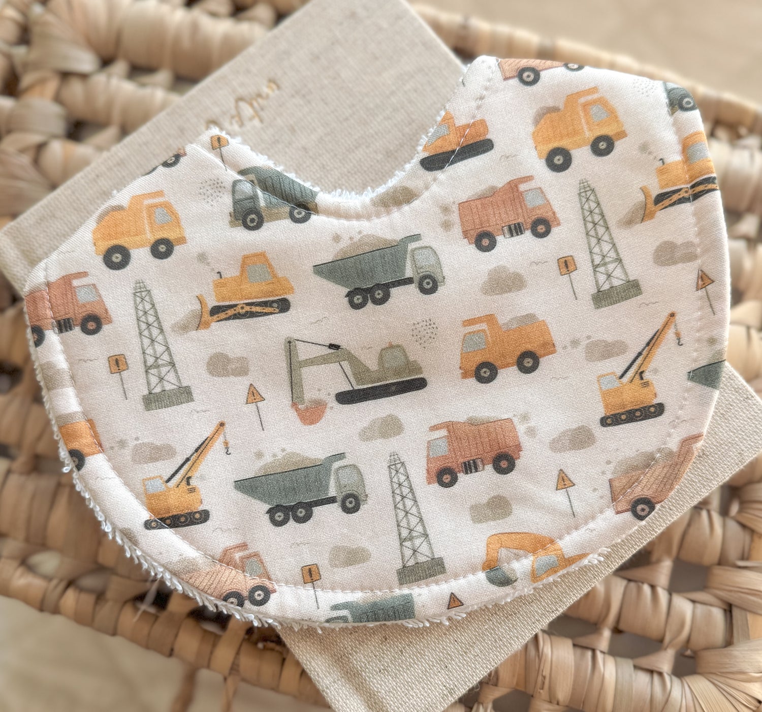 Handmade baby bib featuring a construction-themed print with diggers, cranes, and dump trucks in muted orange, green, and yellow tones. The bib has a curved shape and terry towelling backing, displayed on a neutral linen cloth and woven basket.