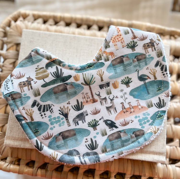 Gender-neutral baby bib with illustrated zebras, giraffes, hippos, birds, and meerkats gathered at desert waterholes; handmade in Australia with soft, absorbent terry towelling backing.