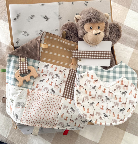 A gift box full with baby gifts including a brown monkey toy comforter, a dark brown check gingham dummy clip, a blue gingham bib, illustrations of jungle animals on another bib and a patchwork taggie blankie with wooden elephant teether. The taggie blankie has mainly light brown, mustard, blue tones with a chocolate brown soft mini fabric on the back. Some of the animals shown in the fabrics are: zebra, giraffe, lion, elephant, monkey, cheetah.