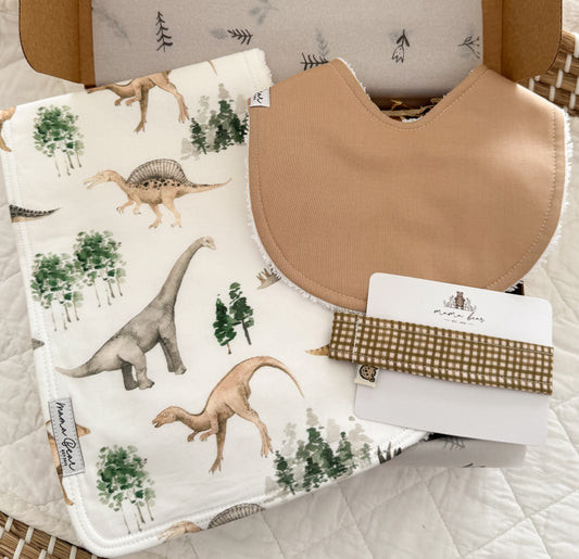 A baby gift set featuring a dinosaur-print burp cloth, a beige bib, and a olive gingham dummy clip, presented in a kraft box.