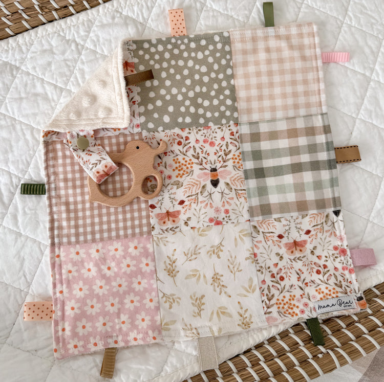 Patchwork quilt style baby taggie with pink florals, polka dot, and checkered patterns. Attached to the taggie is a wooden bee shape. The Blankie includes soft pink, beige and sage green tones.
