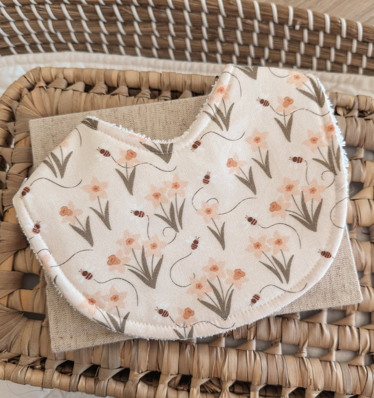 Curved handmade baby bib with a floral and bee pattern printed on cream fabric. The design includes small orange flowers, green leafy stems, and tiny brown bees, styled in a delicate, nature-inspired theme. The bib is placed on a beige linen cloth atop a woven basket surface.