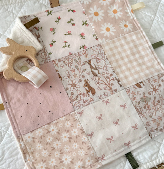 Patchwork baby blanket with floral and animal patterns, accompanied by a wooden teething ring.