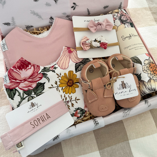 Pink Baby girl gift hamper featuring a solid pale pink bib, another bib with scattered roses and daisy flowers in pinks and mustards, two matching hairbows, a pair of baby shoes with pink piggy faces and a floral swaddle in pink, pale yellow and green tones.
