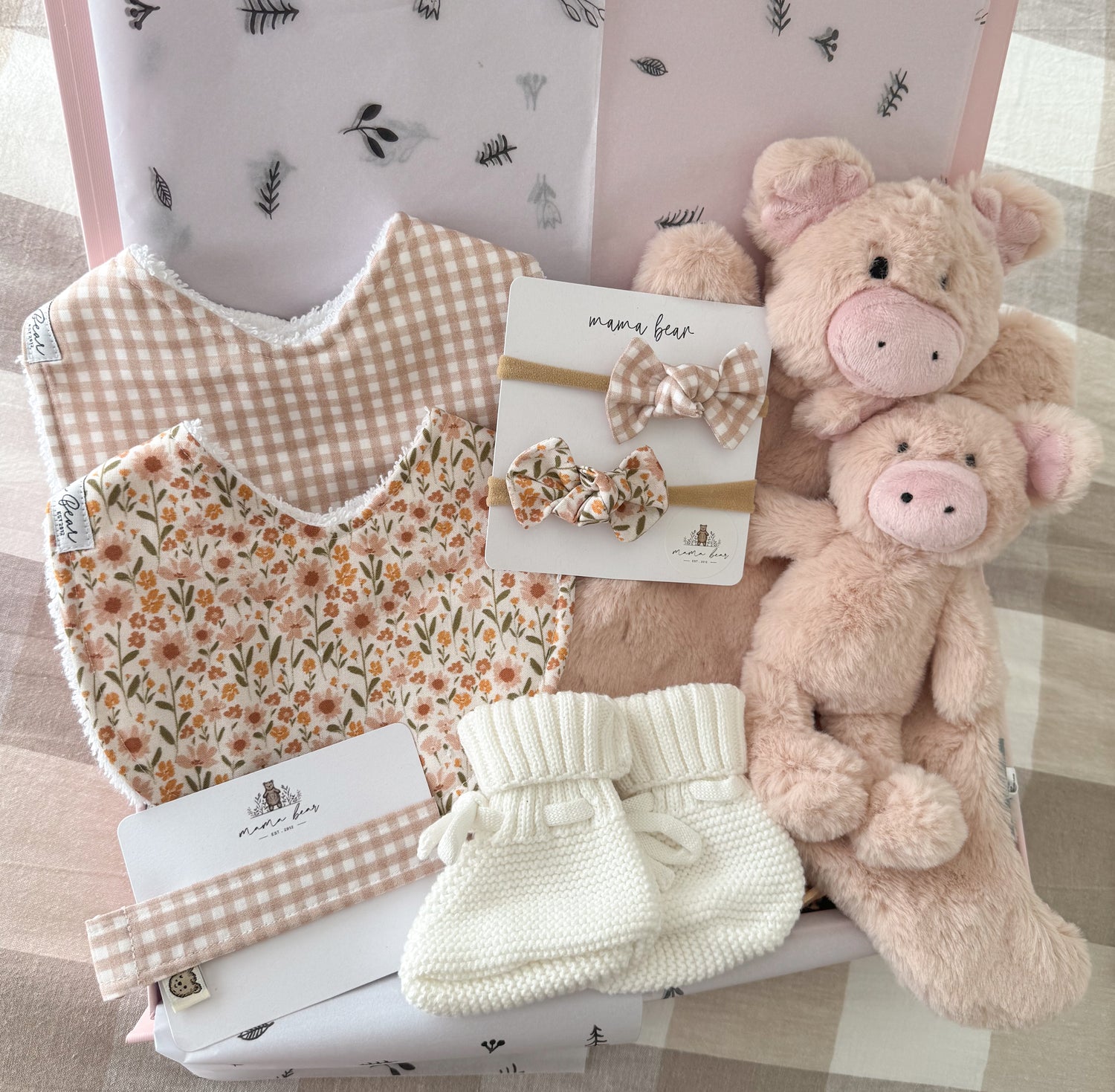 Baby girl pink gift box wirth handmade baby bibs, one in a latte coloured mini gingham check with white background, the other bib is filled with tiny flower stems in colours pink, orange, coral, green. There is two matching hair bows in the same fabric and a dummy clip in the gingham shade. There is a white knitted pair of baby booties and a pale pink soft pig comforter and matching mini pig rattle doll.
