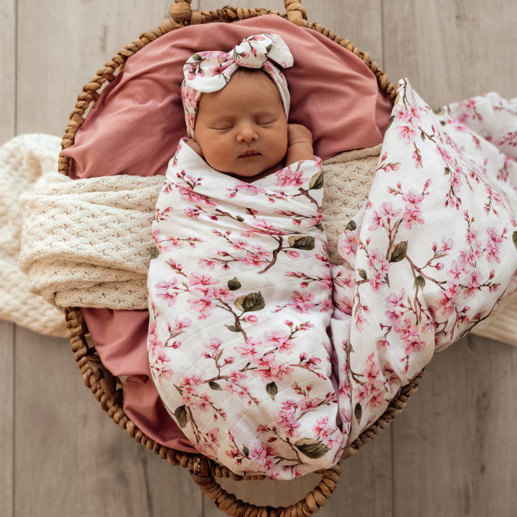 Newborn baby girl wrapped in a pink floral muslin wrap with a matching headband, lying in a wicker basket.