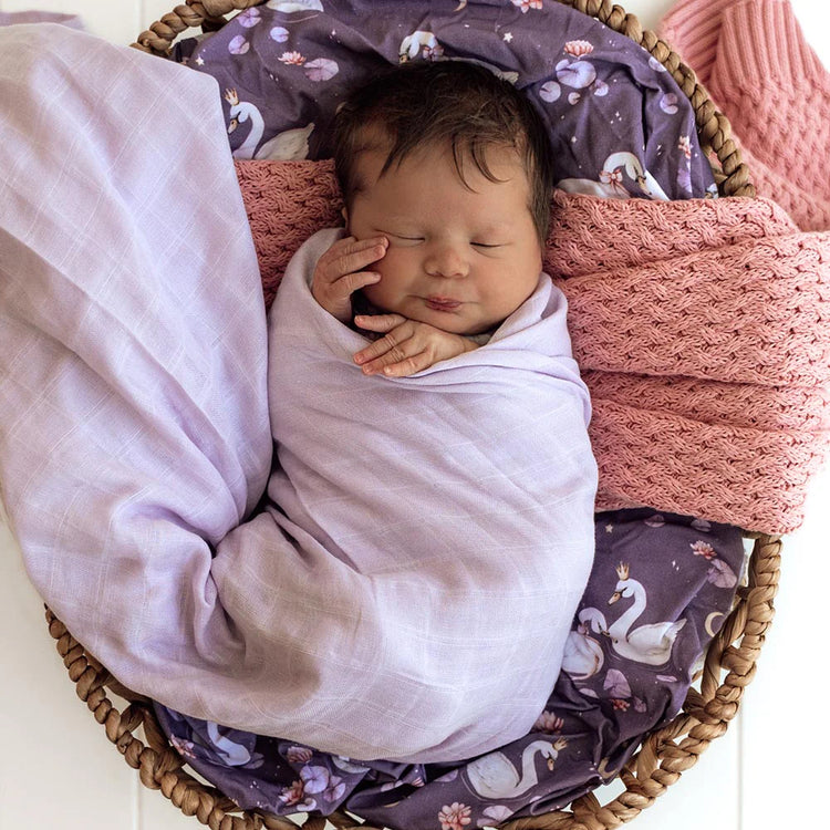 Newborn baby swaddled in a light purple blanket in a wicker basket with pink and purple blankets.