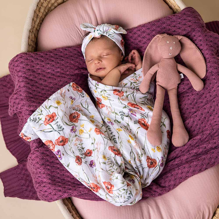 Newborn baby wrapped in a floral blanket with a soft toy, lying on a pink cushion.