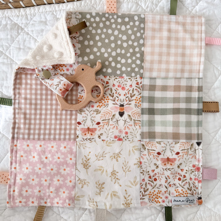 Patchwork quilt style baby taggie with pink florals, polka dot, and checkered patterns. Attached to the taggie is a wooden bee shape. The Blankie includes soft pink, beige and sage green tones.