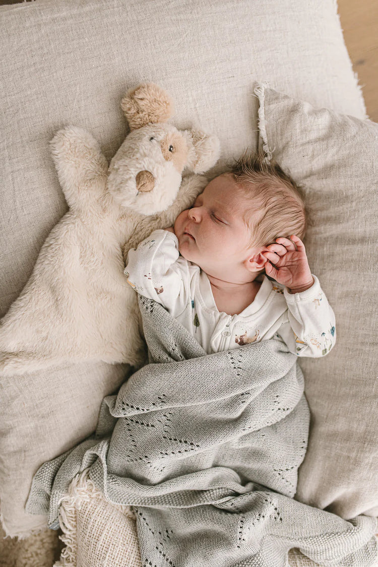Baby asleep on a beige pillow wrapped in a grey blanket. Beside the baby lies a baby toy comforter in a plush light toned fur with a light brown spot on his ear and eye.