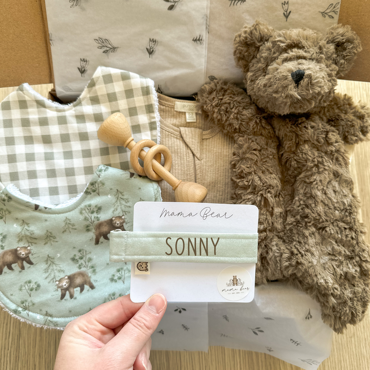 Personalised baby boy gift box with a brown bear comforter, sage dummy clip, two baby bibs (bear print and gingham), wooden rattle, and beige bodysuit. Perfect for baby showers or as a newborn gift hamper in Australia.